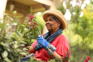A smiling senior woman wearing a straw gardening hat, and gardening gloves holding a pair of gardening pruners and a tall flower stem as she gets ready to cut the stem of the blooming flower.