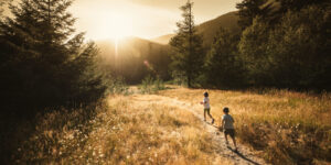 A young girl and a young boy running down a narrow, rocky, dirt mountain trail that is surrounded by dried grass and pine trees on either side at sunrise.