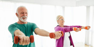 Smiling senior man and smiling senior woman exercising. Both have dumbbells in each hand with their arms raised directly in front of them doing front lateral raises.